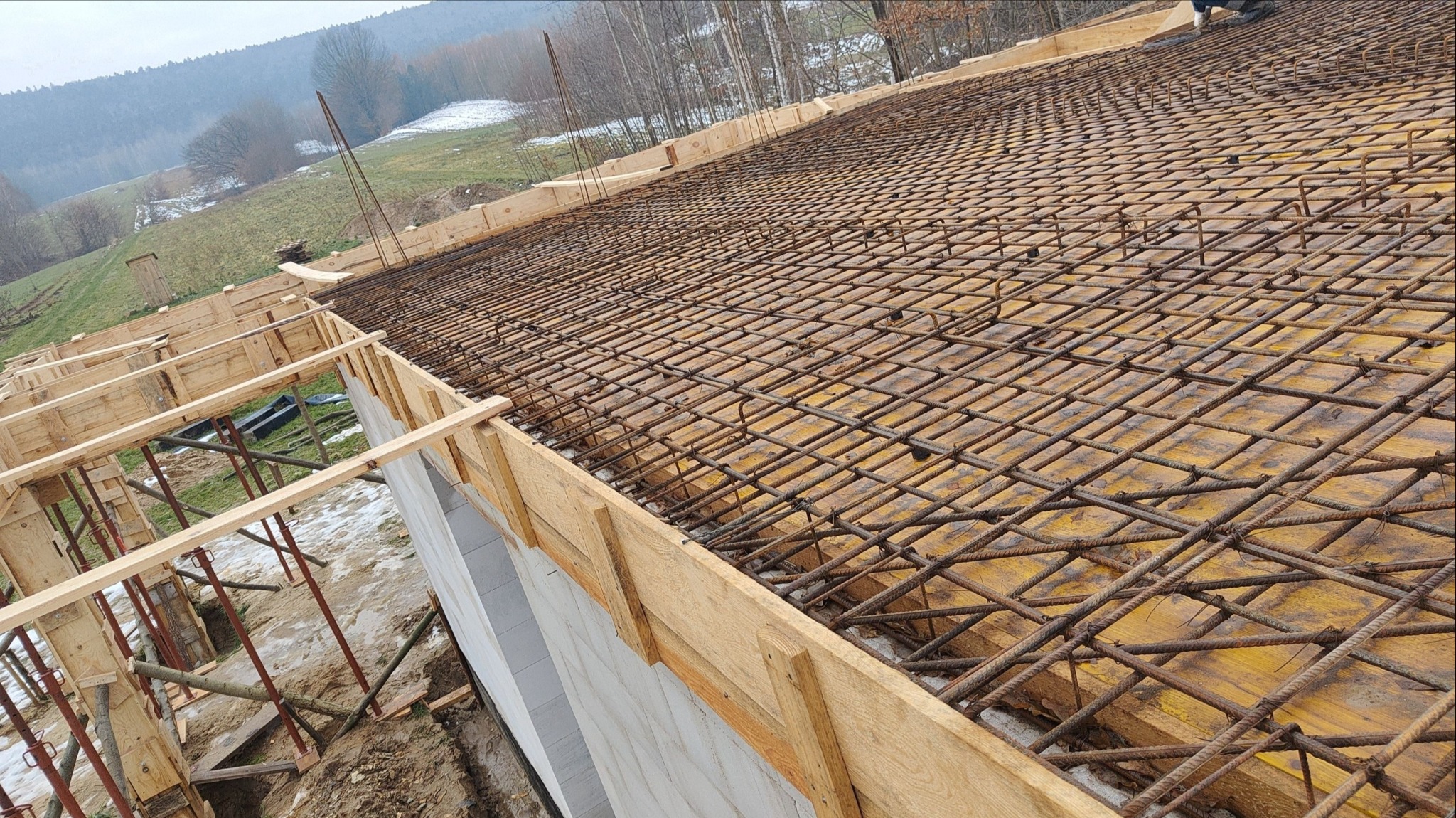 Zbrojenie stropu na deskowaniu, widok z góry. Stalowe pręty tworzą siatkę na żółtej powierzchni, w tle widać zalesione wzgórza i fragment konstrukcji szalunkowej.