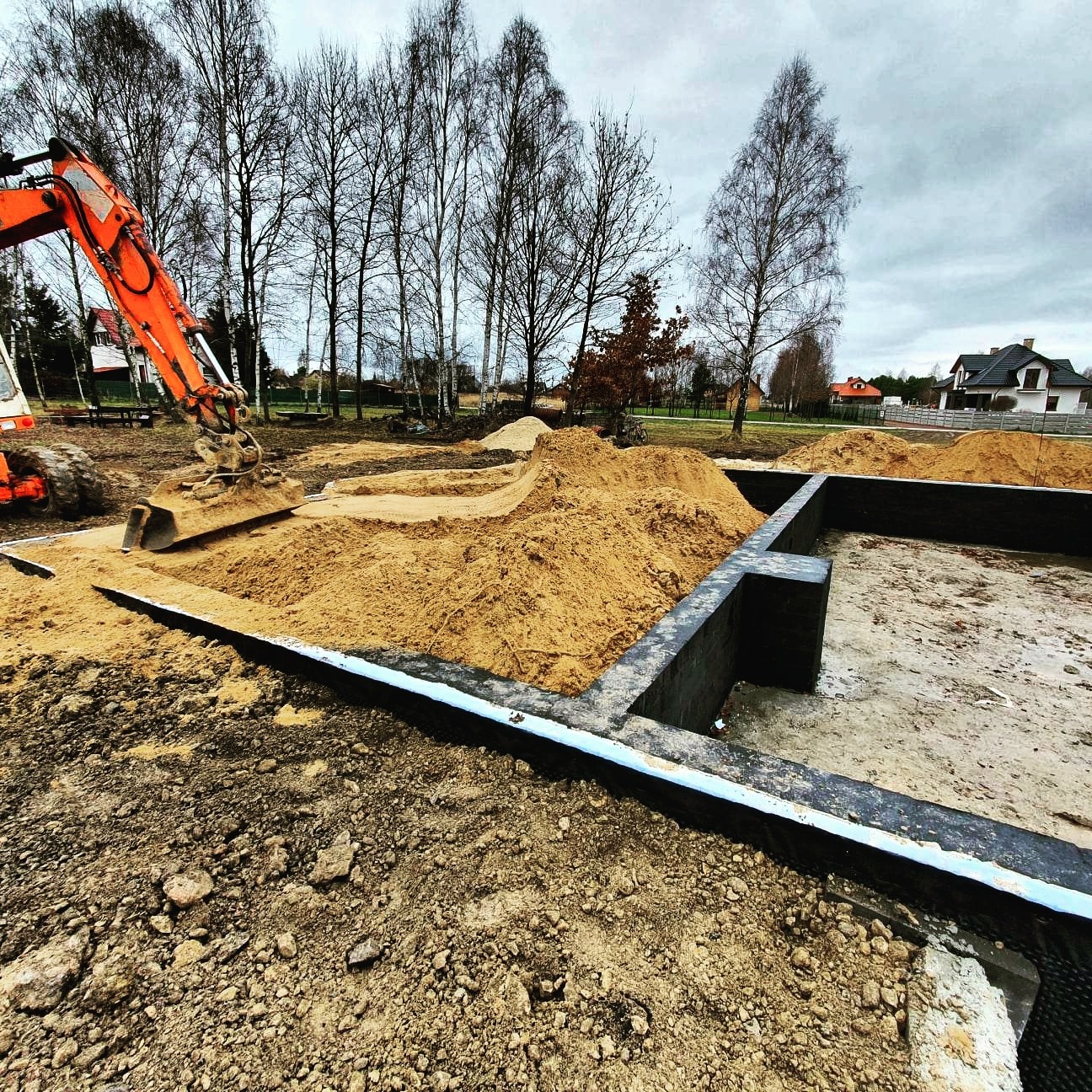 Fundamenty domu w trakcie budowy: wykop, szalunek i piasek. Pomarańczowa koparka w tle. Widok na plac budowy w pochmurny dzień.