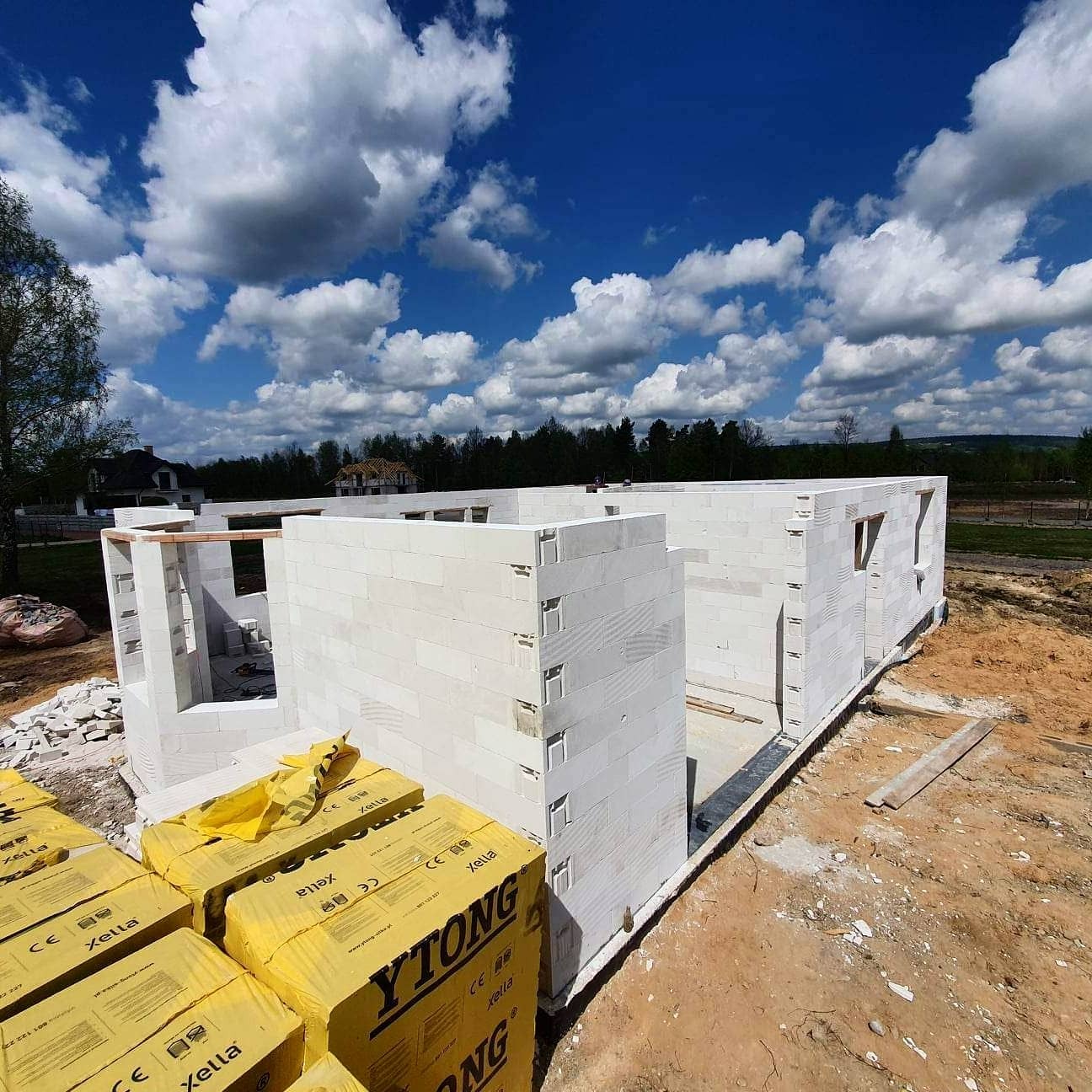 Wznoszenie ścian domu z białych bloczków Ytong. Częściowo wymurowane ściany budynku na tle błękitnego nieba z białymi chmurami. Na pierwszym planie paleta bloczków.