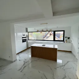 Nowoczesna kuchnia z białymi, lakierowanymi szafkami, drewnianą wyspą i marmurową podłogą, oświetlona złotą lampą wiszącą i światłem dziennym z dużego okna z widokiem na domy.