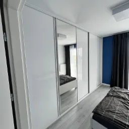 Biała szafa wnękowa z przesuwnymi drzwiami z lustrami w sypialni z szarą podłogą i niebieską ścianą w tle.