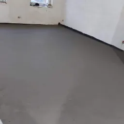 Świeżo wylana, gładka posadzka cementowa w pomieszczeniu z oknami, zabezpieczona czarną folią przy ścianach.