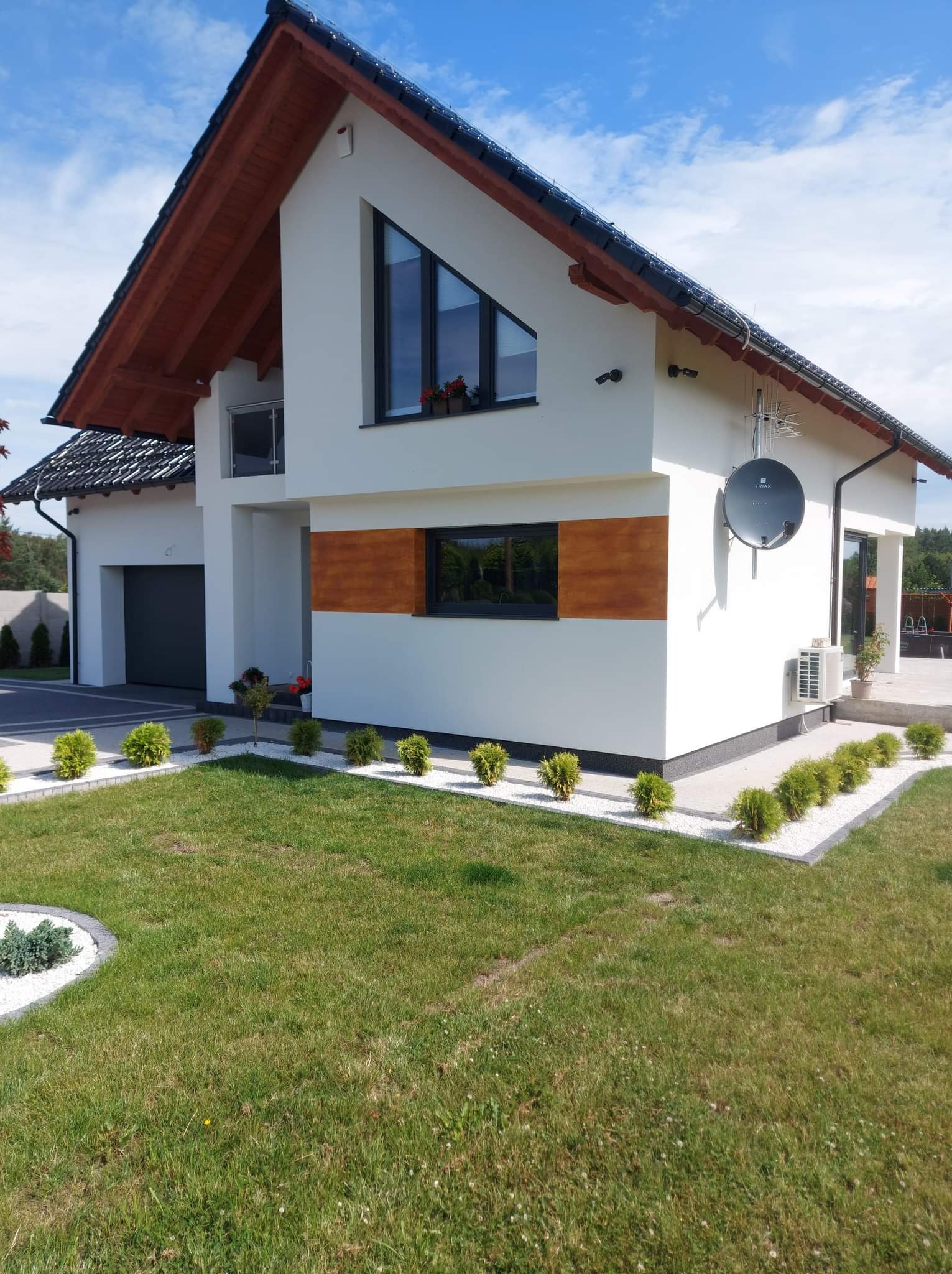 Biały dom jednorodzinny z drewnianymi akcentami, czarną dachówką i zadbanym trawnikiem z ozdobnymi krzewami i białym żwirem. Widoczna antena satelitarna i klimatyzator na elewacji.