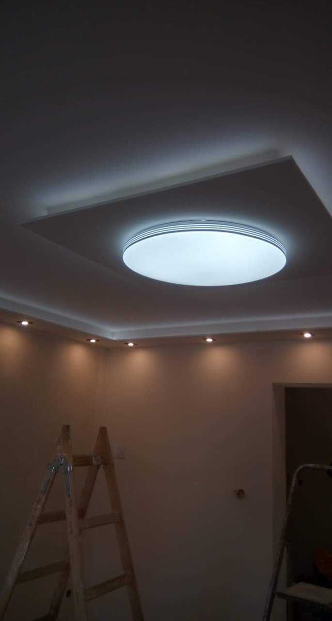 Nowoczesne oświetlenie sufitowe z okrągłą lampą i punktowymi światłami, widoczne drabiny malarskie w pomieszczeniu w trakcie remontu.