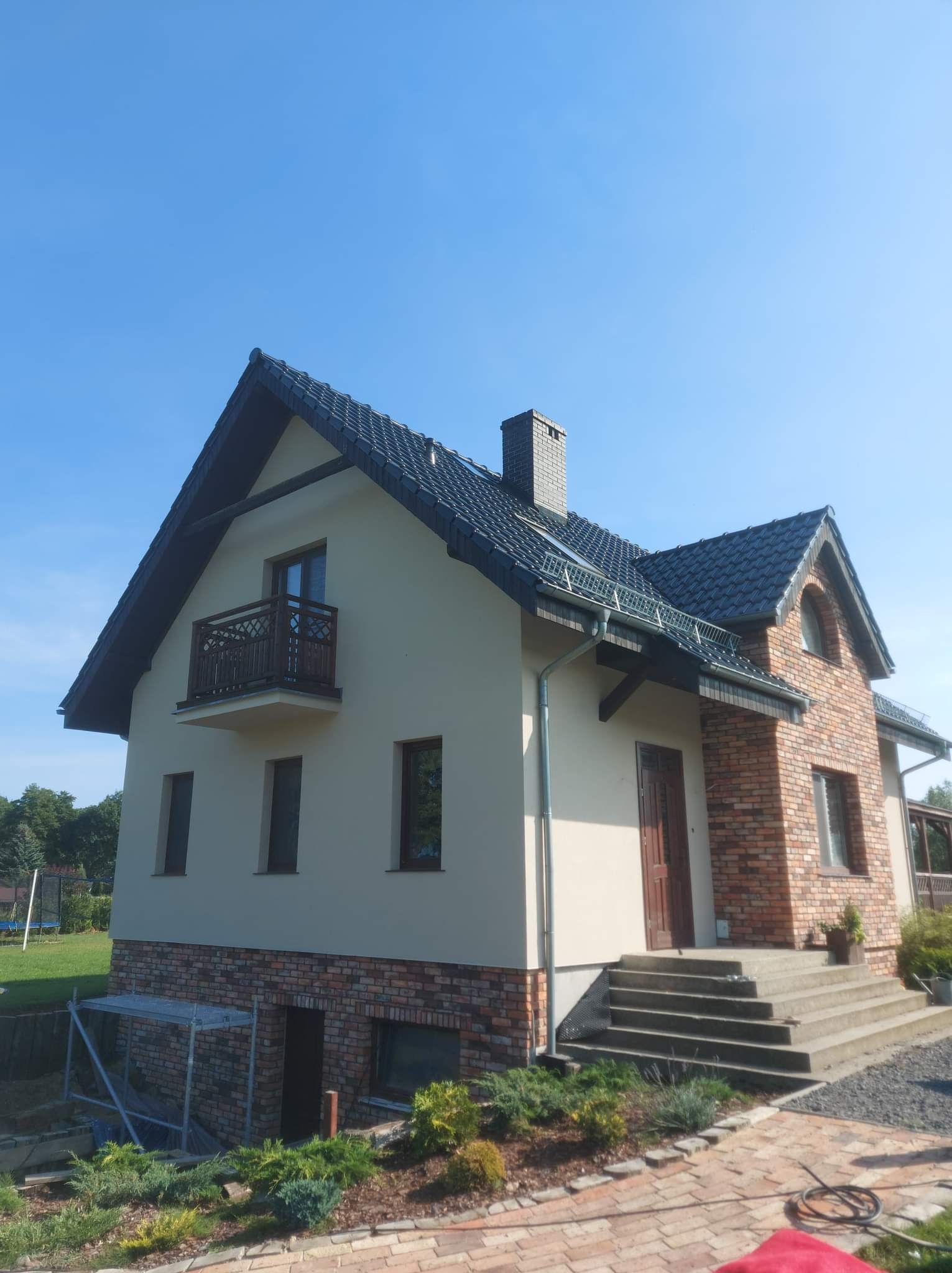 Dwukondygnacyjny dom z beżową elewacją i ceglanym wykończeniem fragmentu ściany oraz fundamentów, widoczne rusztowanie przy dolnej części budynku, dachówka w kolorze grafitowym, jasne niebo.