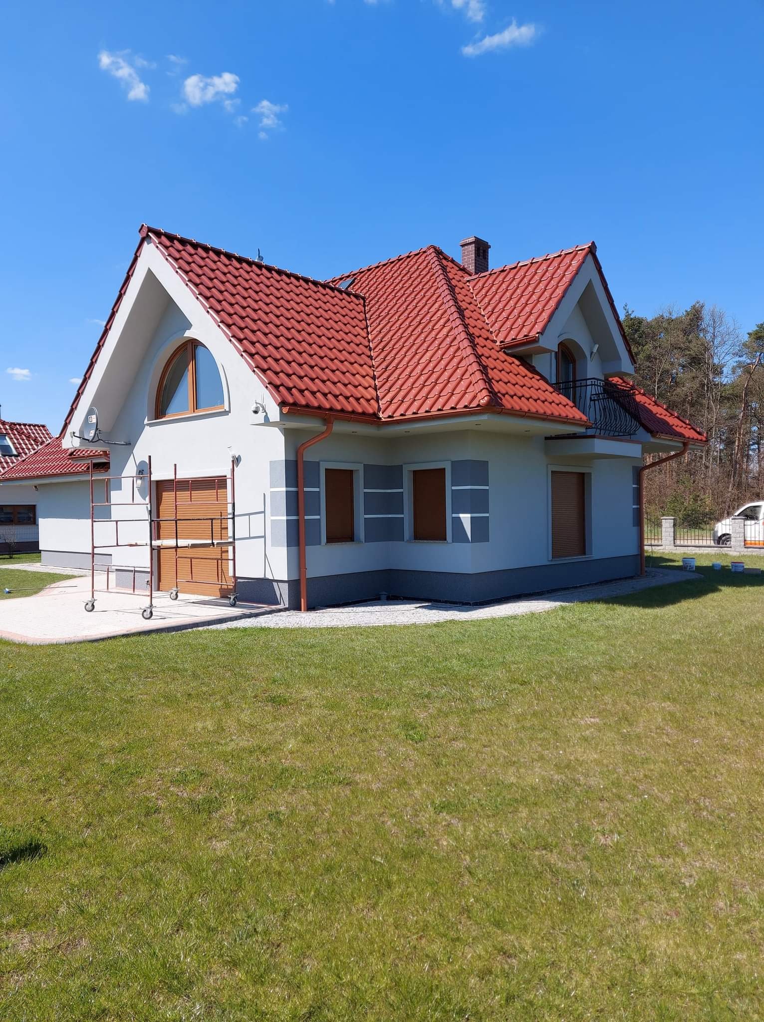 Ocieplony dom jednorodzinny z czerwoną dachówką i szarymi akcentami, częściowo zasłonięty rusztowaniem na tle błękitnego nieba.