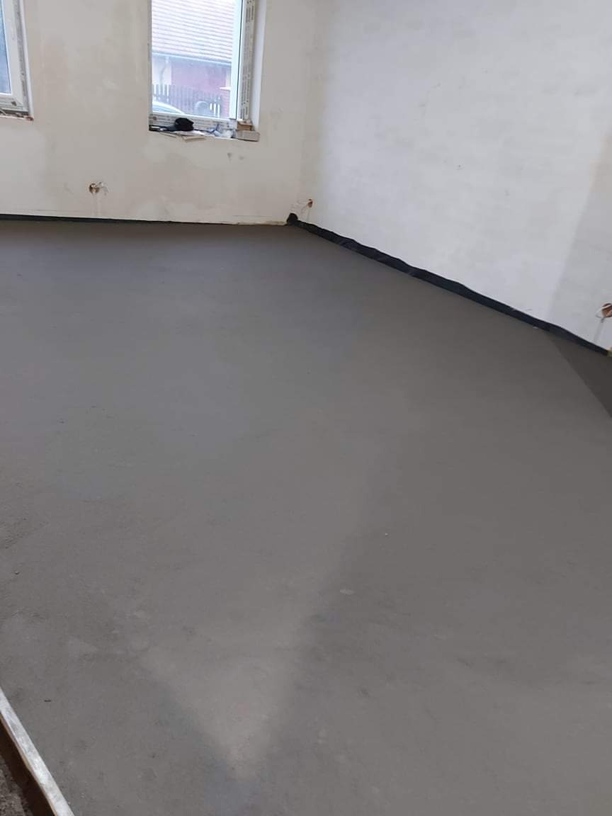 Świeżo wylana, gładka posadzka cementowa w pomieszczeniu z oknami, zabezpieczona czarną folią przy ścianach.