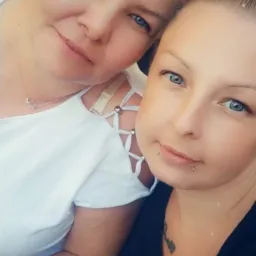 Portret dwóch uśmiechniętych kobiet z jasnymi oczami, jedna z krótkimi siwymi włosami i naszyjnikiem z gwiazdą, druga z tatuażem na szyi i piercingiem w dolnej wardze.