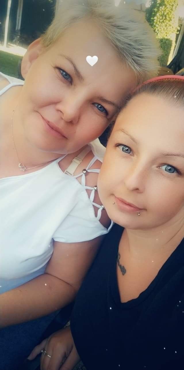 Portret dwóch uśmiechniętych kobiet z jasnymi oczami, jedna z krótkimi siwymi włosami i naszyjnikiem z gwiazdą, druga z tatuażem na szyi i piercingiem w dolnej wardze.