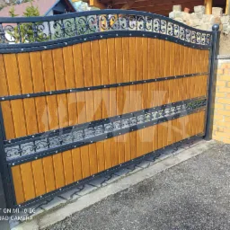 Zamykana brama wjazdowa z drewnianymi panelami w kolorze jasnego dębu, ozdobiona czarnymi, kutymi elementami i nitami, zamontowana na słupku z cegły klinkierowej z numerem posesji.