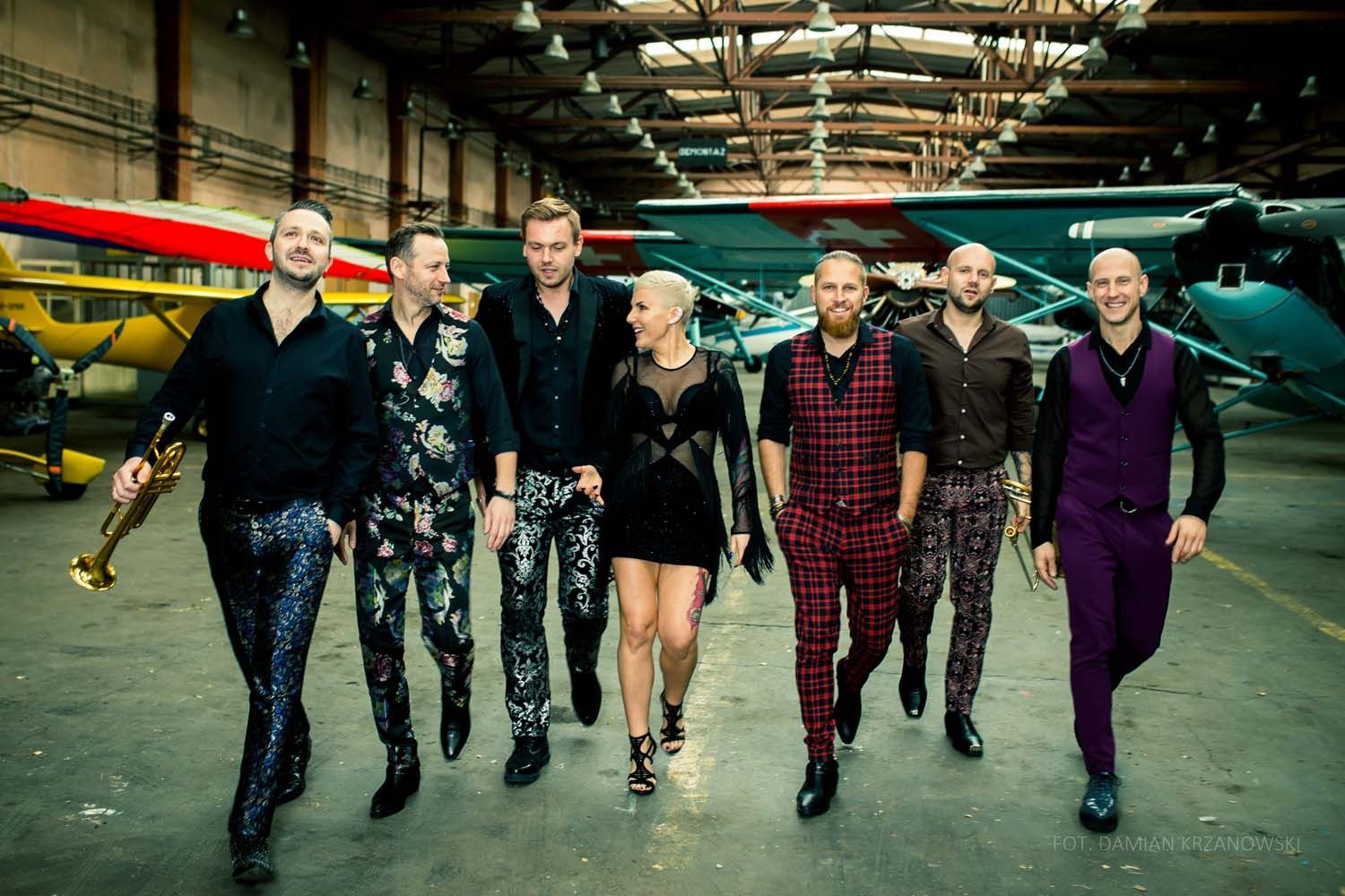 Zespół muzyczny w ekstrawaganckich strojach pozuje na tle hangarów z samolotami.