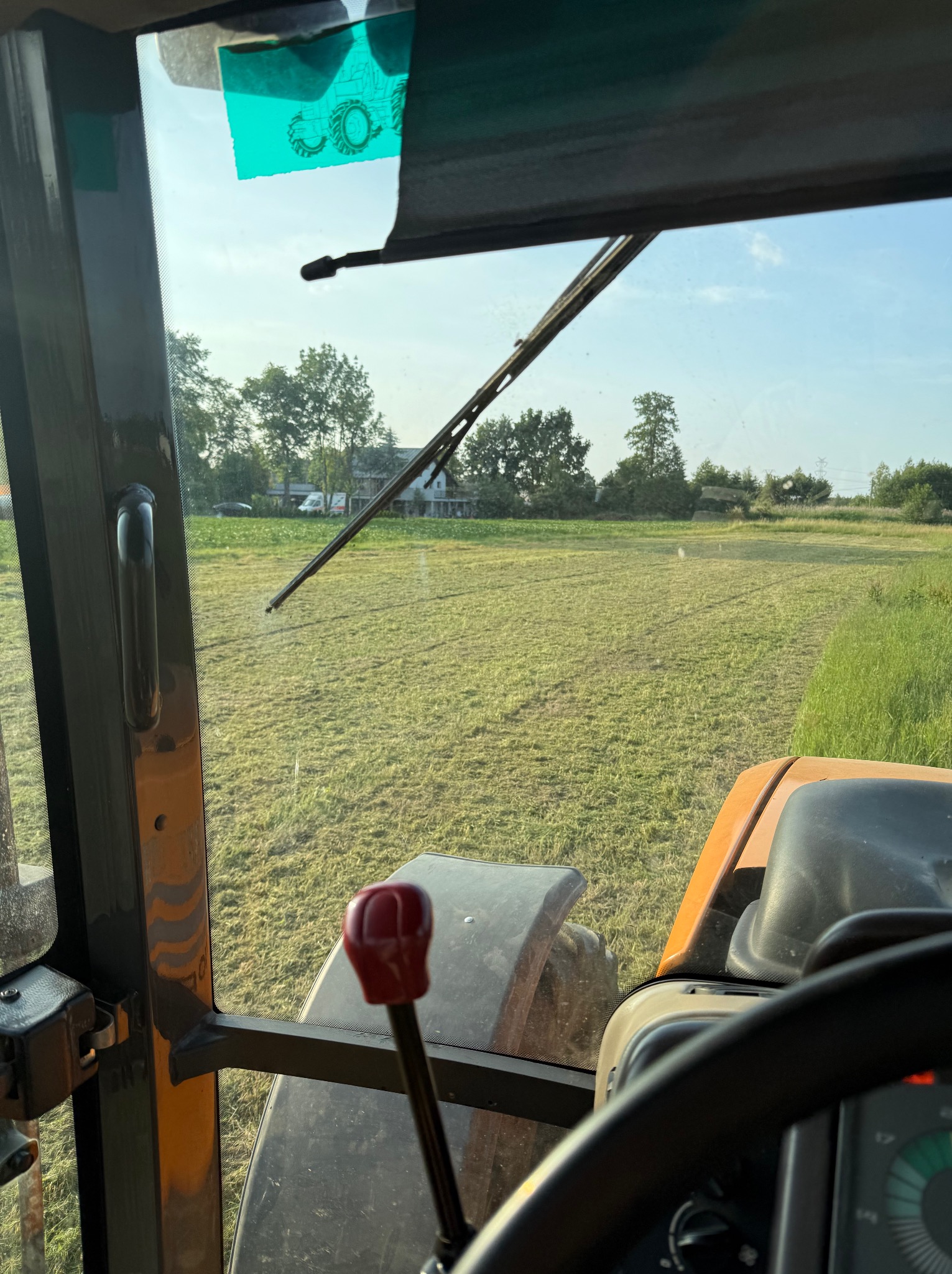 Widok z kabiny traktora na pole po koszeniu trawy w słoneczny dzień. Świeżo skoszona trawa, widoczne drzewa w oddali, szyba z naklejką traktora.