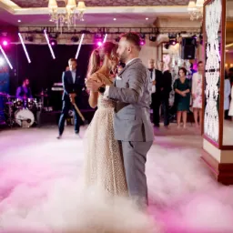 Para młoda tańczy pierwszy taniec na parkiecie spowitym sztuczną mgłą, w tle zespół muzyczny i goście, oświetlenie sceniczne w odcieniach różu i fioletu.