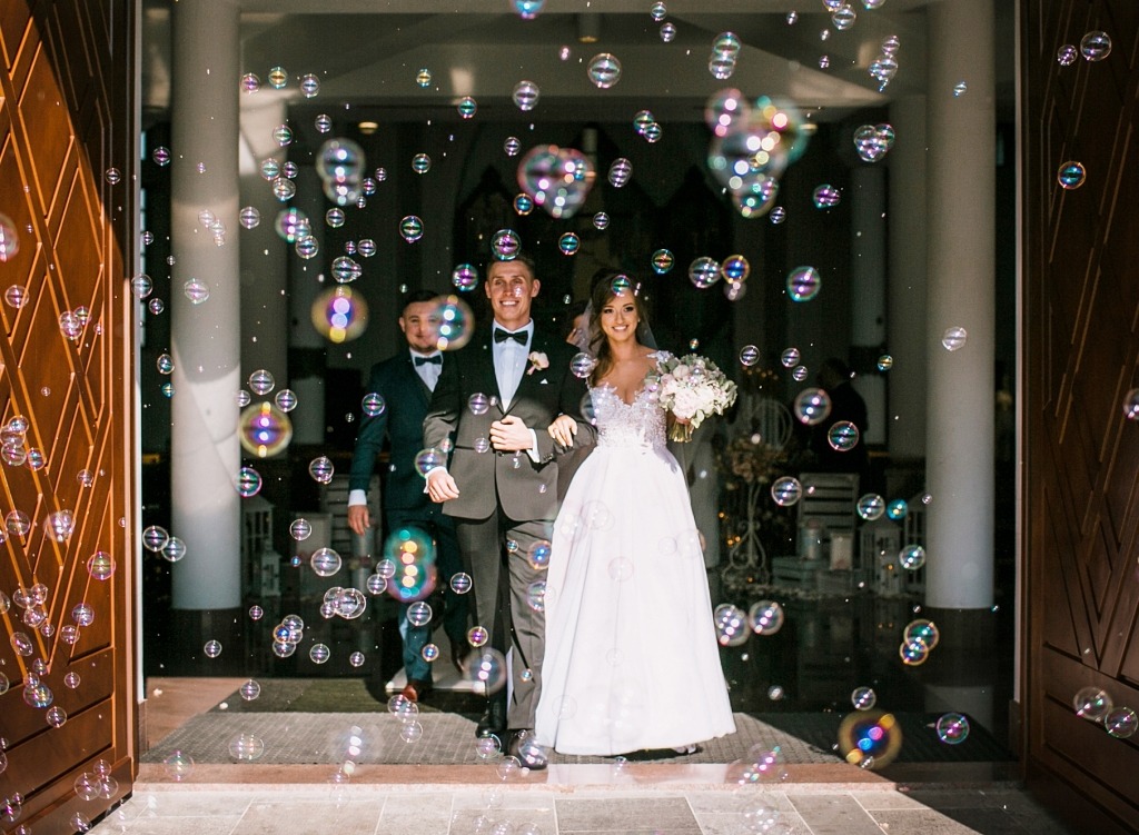 Para młoda wychodzi z kościoła, otoczona bańkami mydlanymi, w tle widoczny druh w garniturze.