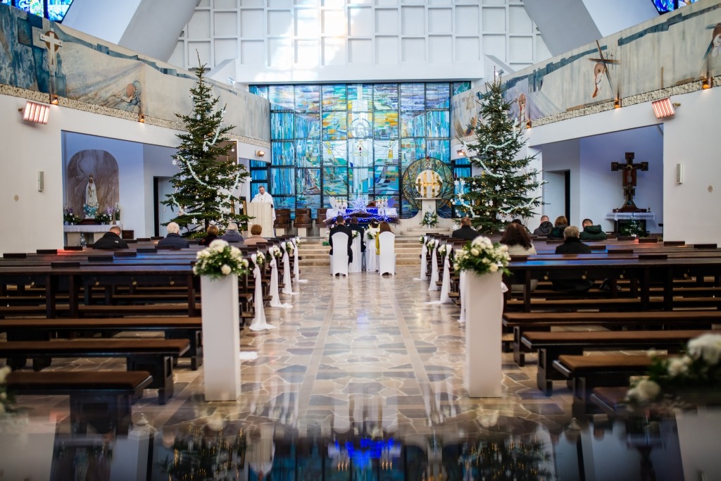 Dekoracja kościoła na ślub: białe kwiaty w kompozycjach na kolumnach, tiulowe wstążki zdobiące ławki, choinki po bokach ołtarza, para młoda w oddali.