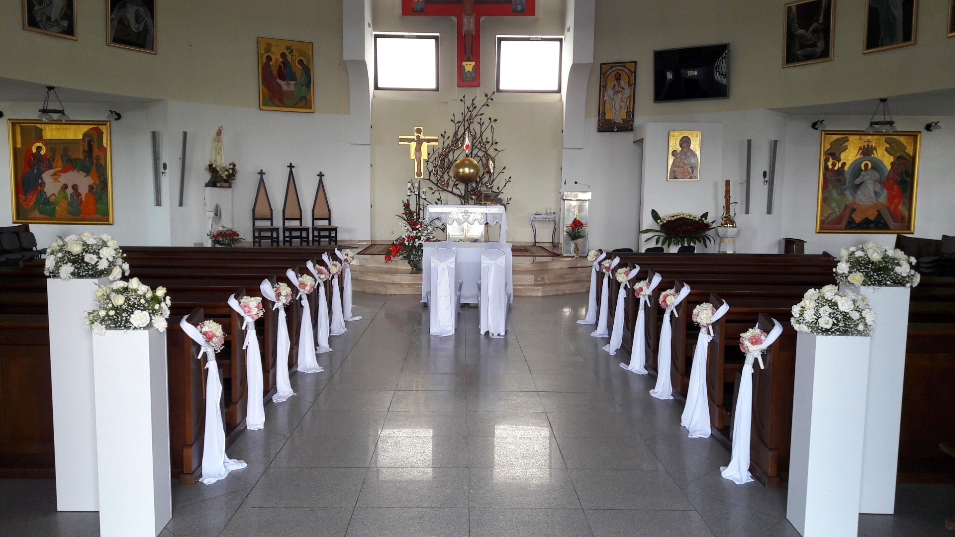 Uroczystość ślubna w kościele: dekoracja ławek białymi wstążkami i kwiatami, białe kolumny z kwiatowymi kompozycjami przy wejściu, ozdobiony ołtarz z białymi pokrowcami na krzesłach.