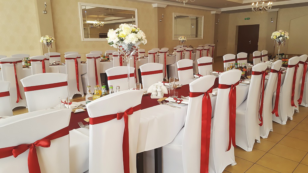 Uroczysta sala bankietowa z długimi stołami nakrytymi białymi obrusami i bordowymi bieżnikami, krzesła w białych pokrowcach ozdobione czerwonymi wstążkami, na stołach dekoracje kwiatowe w wysokich...