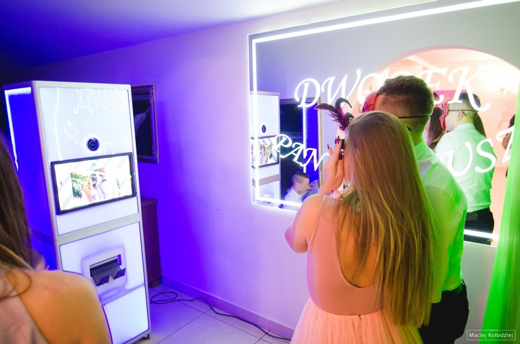 Goście weselni w Żółtańce-Kolonia korzystają z fotobudki z neonowym napisem, robiąc sobie zdjęcia z rekwizytami.