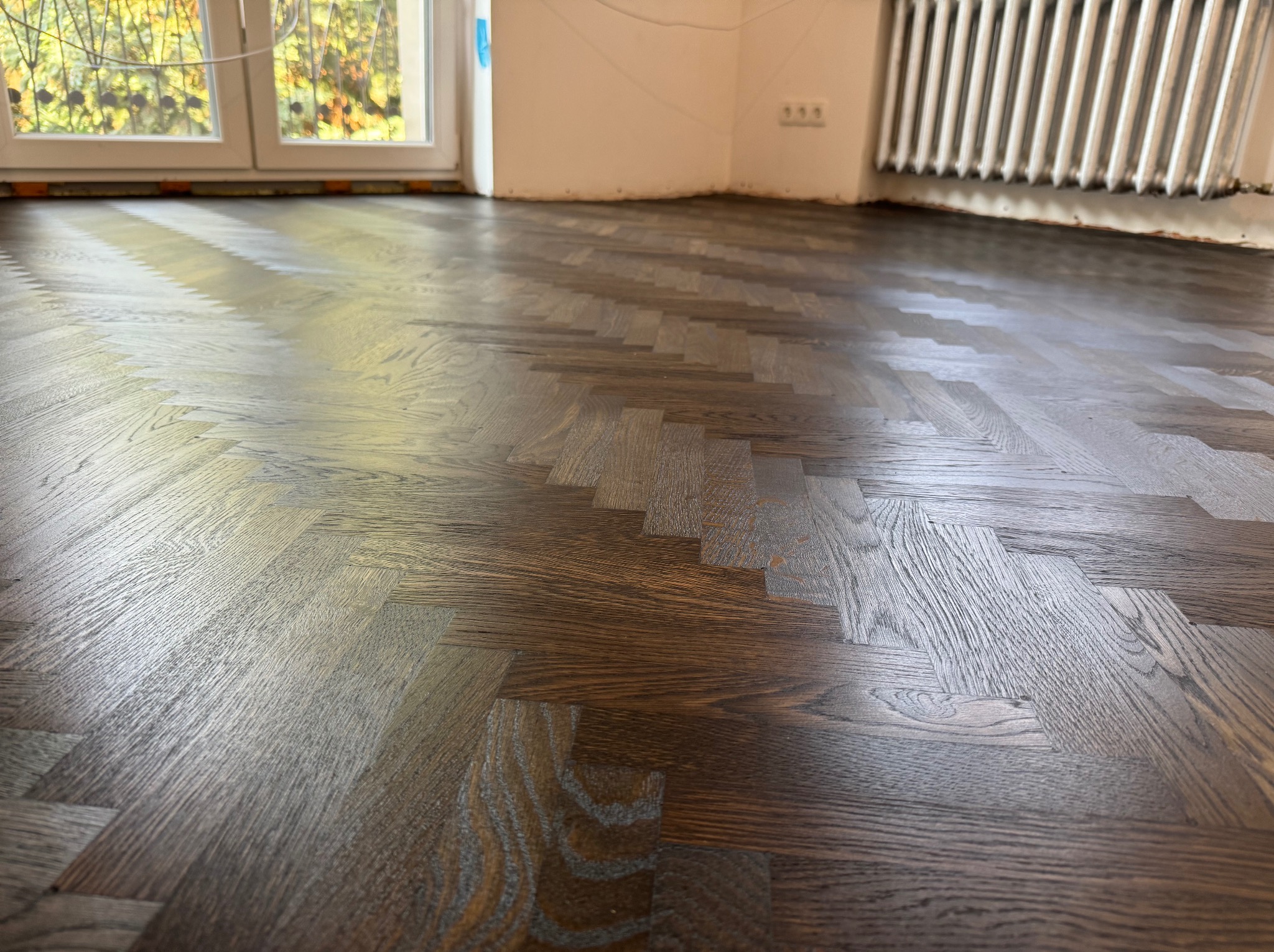 Ciemna, lakierowana podłoga parkietowa ułożona w jodełkę, widoczna w jasnym pomieszczeniu z oknem i grzejnikiem.