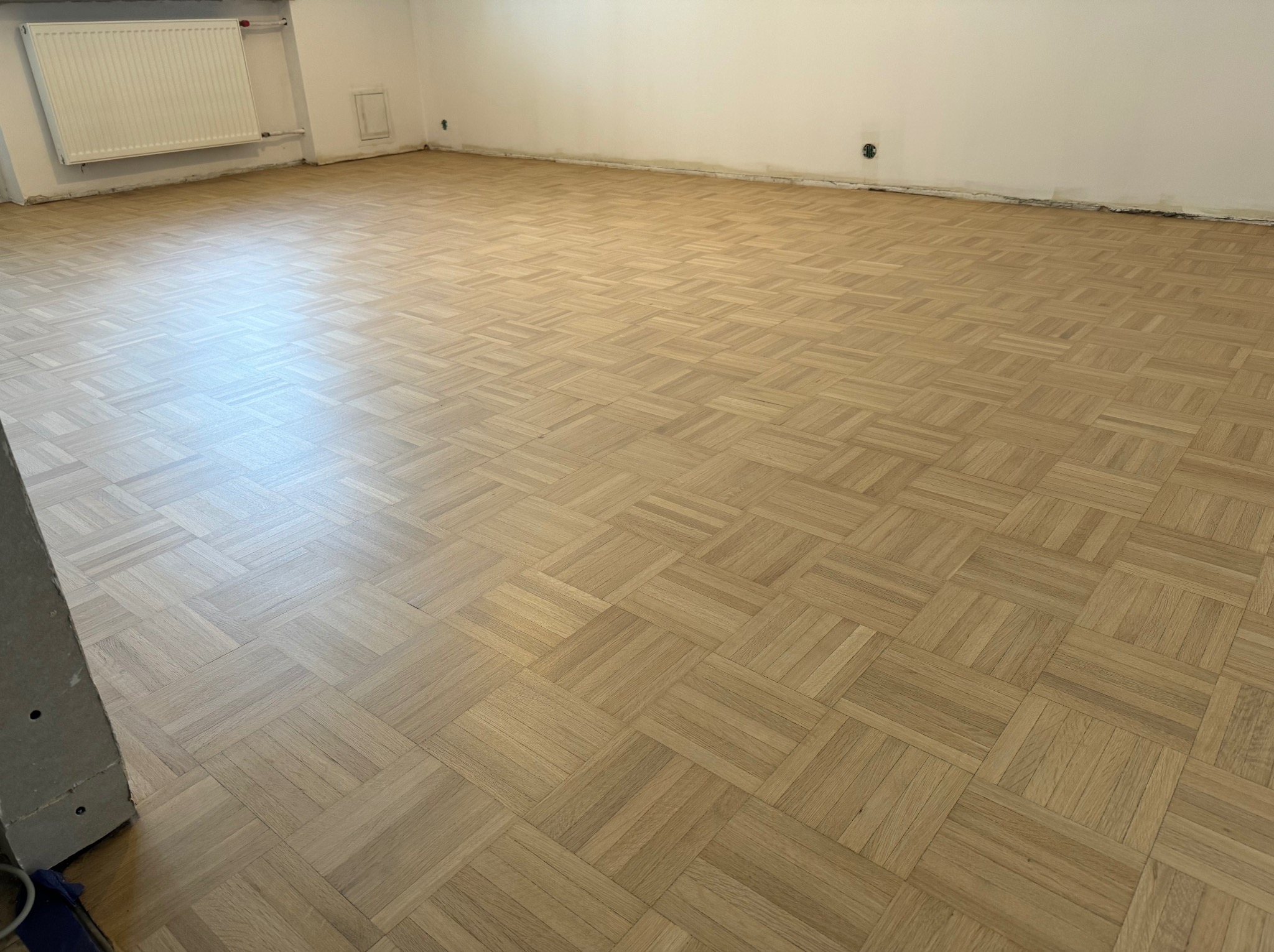 Parkiet ułożony w jodełkę kwadratową, po renowacji, widoczne ślady po listwach przypodłogowych na ścianie, grzejnik na ścianie w tle.
