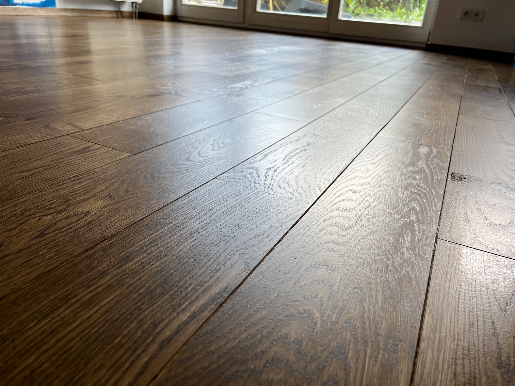 Błyszcząca, odnowiona podłoga z ciemnego drewna, z widocznymi słojami i odbiciami światła, położona w kierunku drzwi balkonowych.