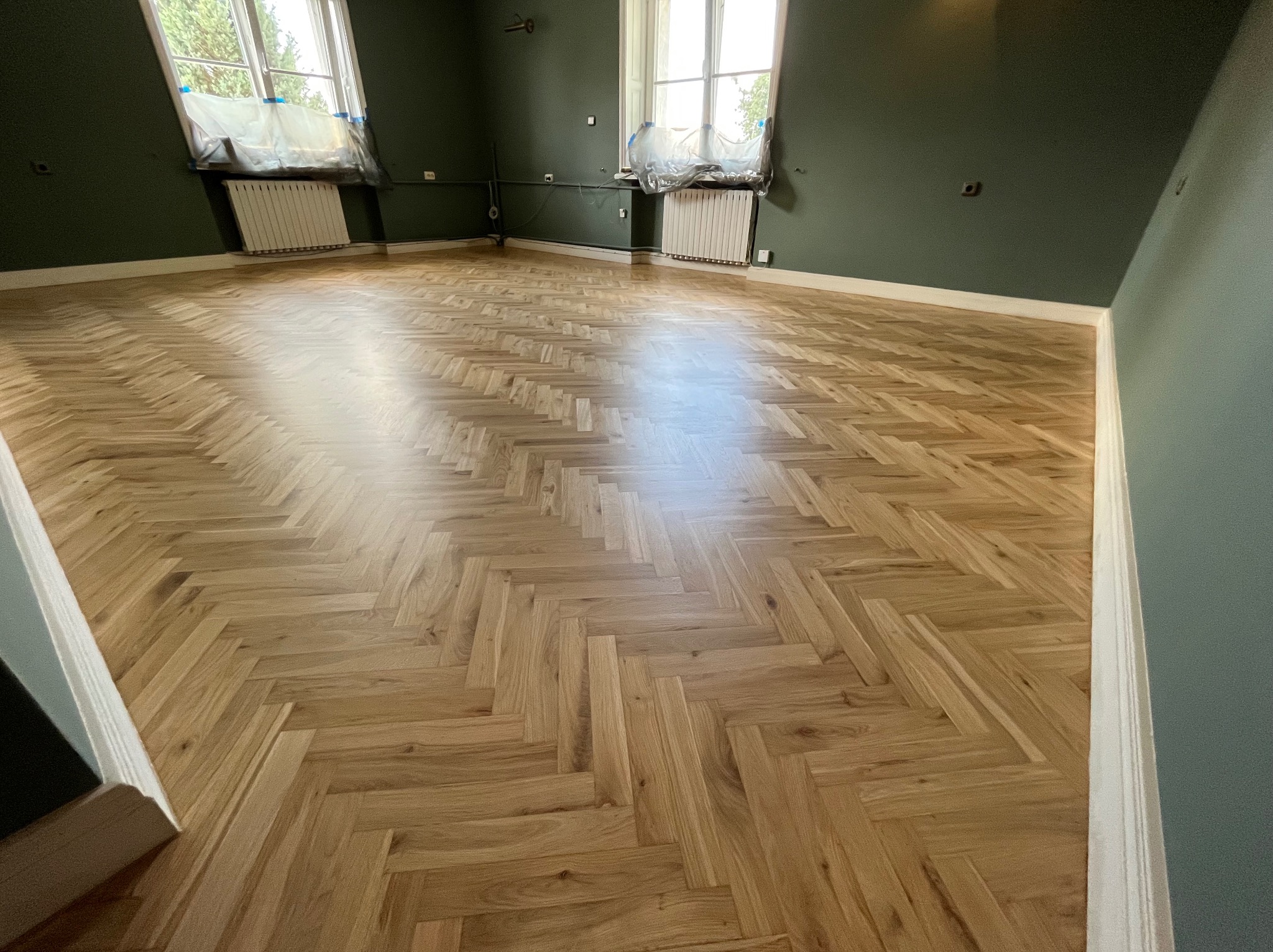 Parkiet ułożony w jodełkę w przestronnym pokoju z ciemnozielonymi ścianami i oknami zabezpieczonymi folią malarską.