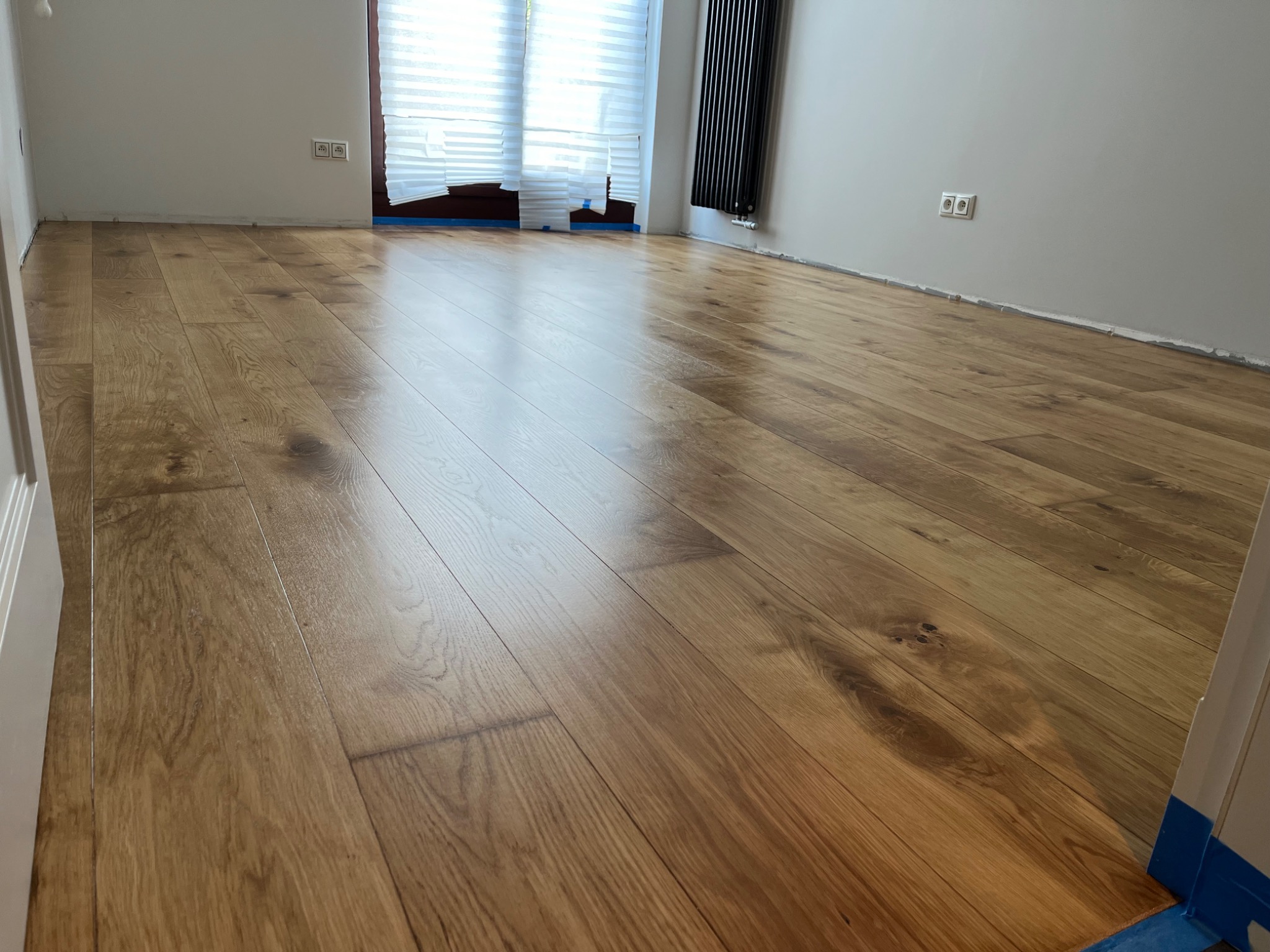Wycyklinowana podłoga parkietowa z widocznymi detalami słojów drewna, zabezpieczona taśmą malarską przy ścianach i ościeżnicy, w pomieszczeniu z grzejnikiem żeberkowym i zasłoniętym oknem.