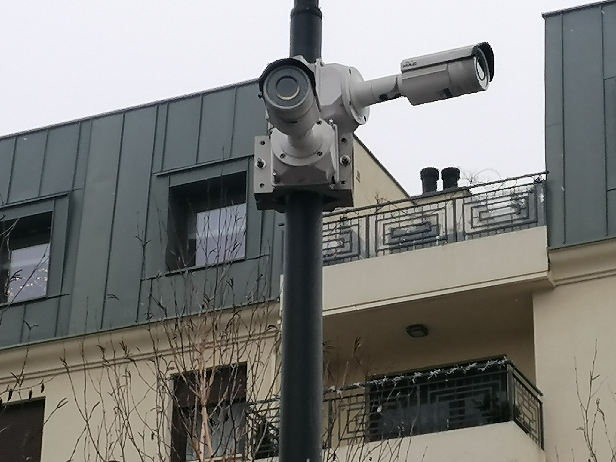 Dwie kamery monitoringu, jedna obrotowa, zamontowane na słupie na tle budynku mieszkalnego z balkonami i szarą blachą na dachu.