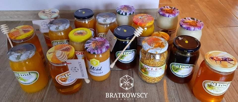 Różne rodzaje miodów w słoikach, z drewnianymi nabierakami, etykiety z logo firmy Bratkowscy, ułożone na drewnianym blacie.