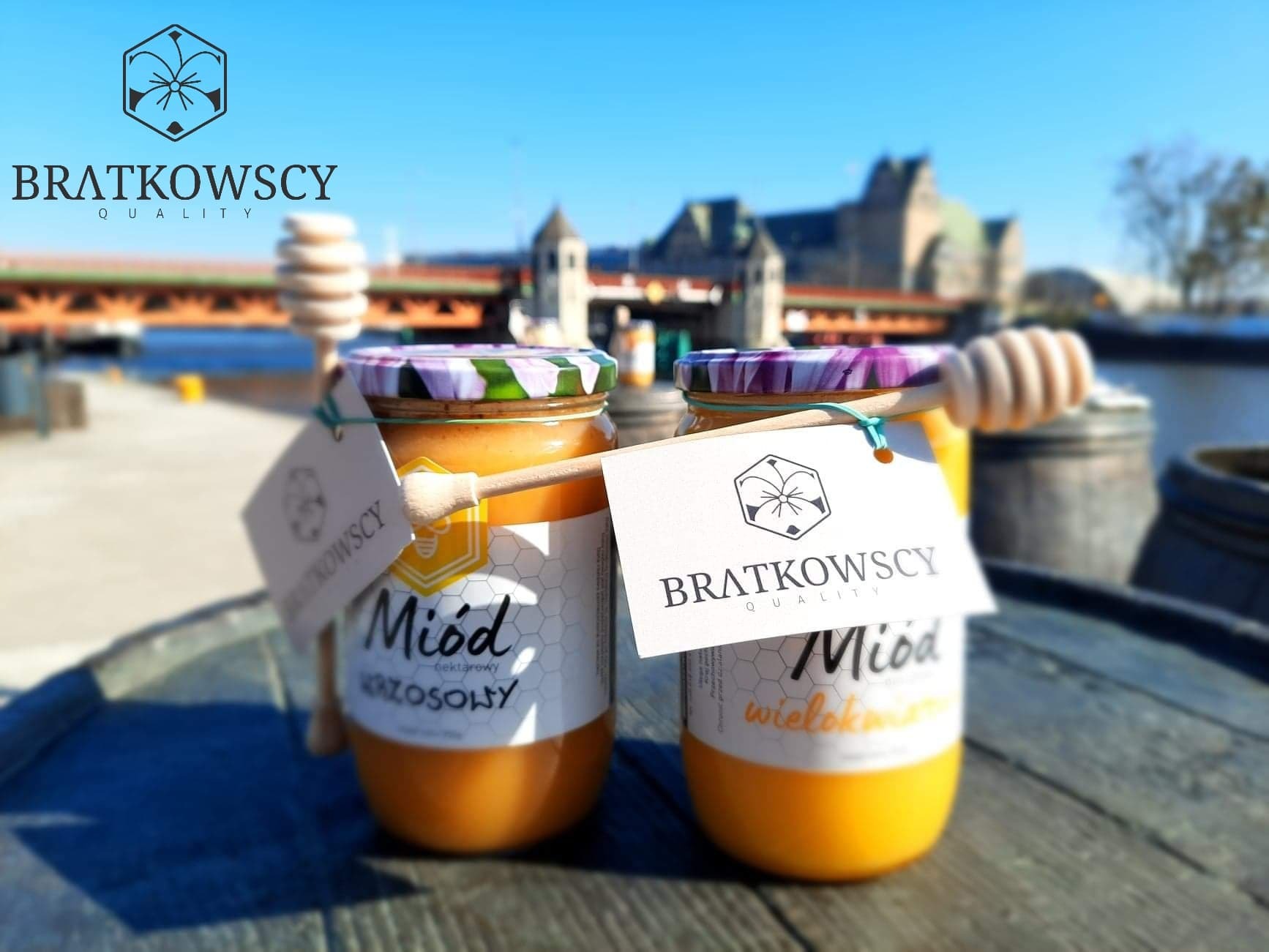 Dwa słoiki z miodem nektarowym i wielokwiatowym marki Bratkowscy, z drewnianymi nabierakami, na tle miejskiego krajobrazu z rzeką i mostem.