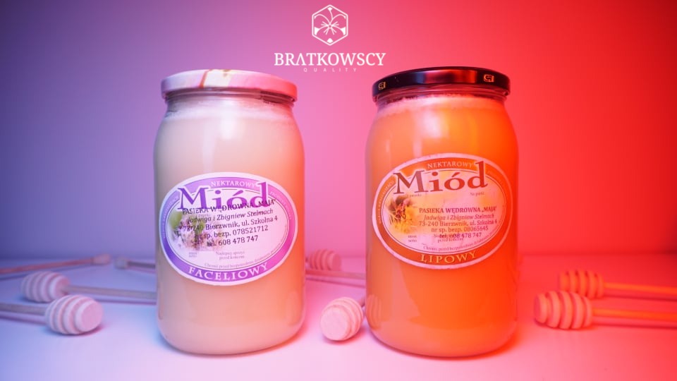 Dwa słoiki miodu nektarowego: faceliowy o jasnej barwie i lipowy o barwie bursztynowej, oba z etykietami Pasieki Wędrownej, na białym tle z drewnianymi nabierakami do miodu, oświetlone światłem...