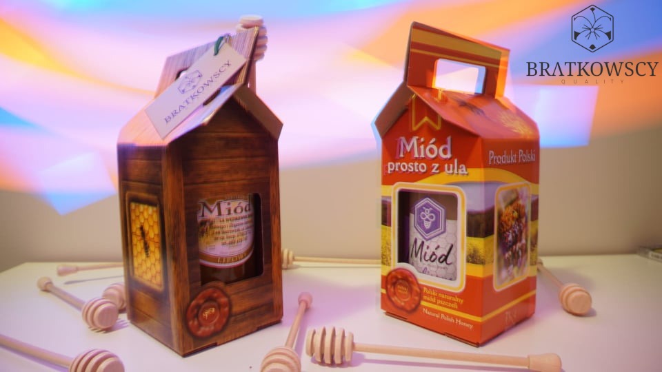 Dwa opakowania miodu marki Bratkowscy, jedno w drewnianym pudełku z okienkiem w kształcie plastra miodu, drugie w pomarańczowym kartoniku, otoczone drewnianymi nabierakami do miodu.