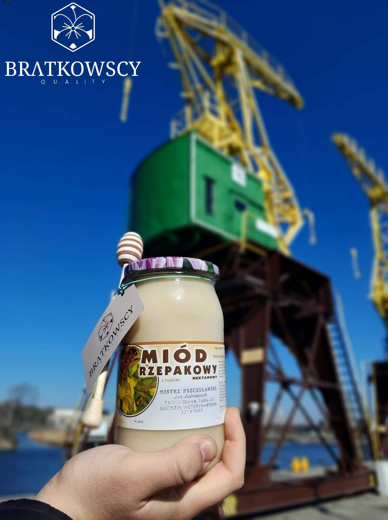 Słoik rzepakowego nektaru z pasieki Bratkowscy, trzymany w dłoni na tle portowych dźwigów i błękitnego nieba.