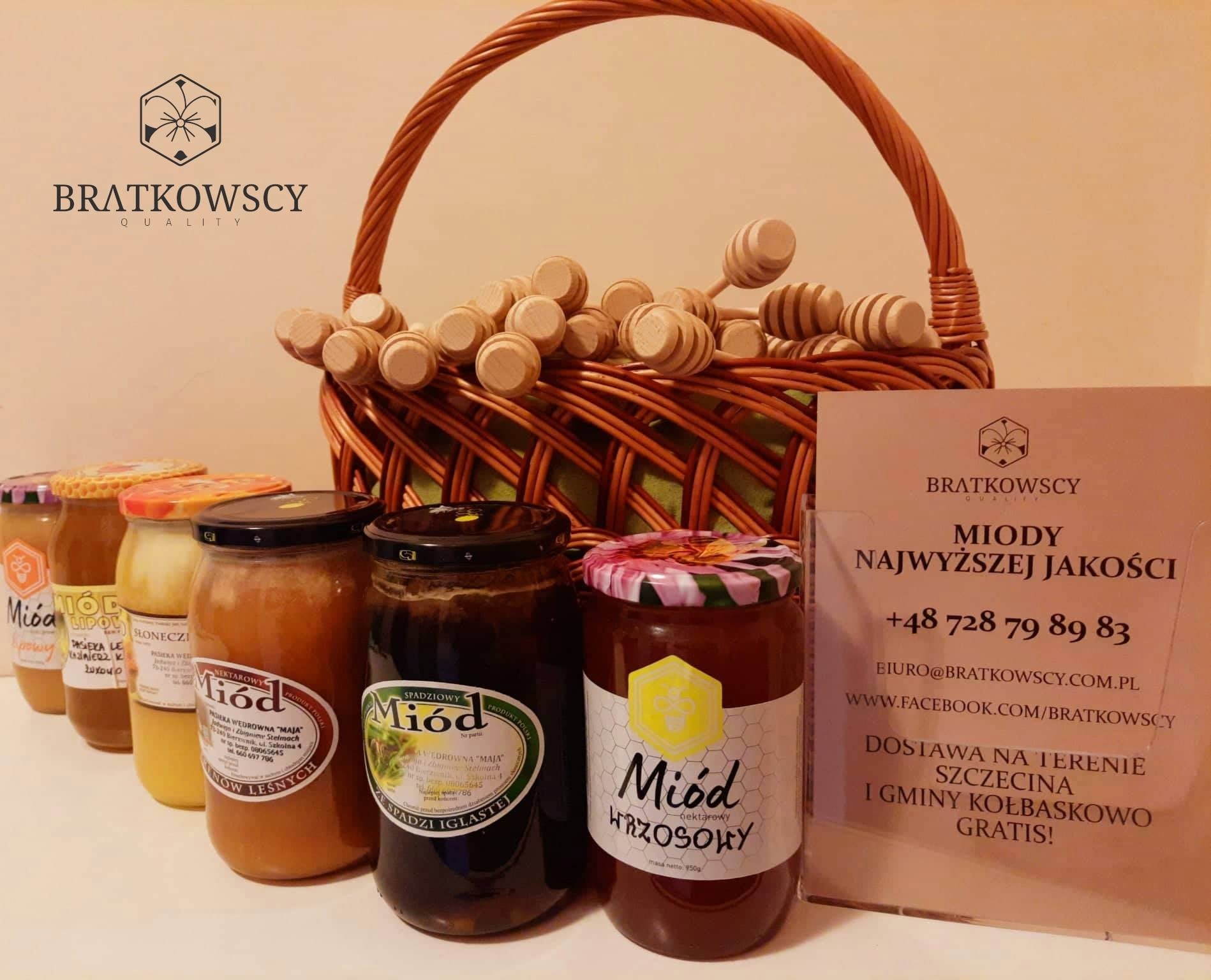 Słoiki z różnymi rodzajami miodu, ustawione w rzędzie przed wiklinowym koszem wypełnionym drewnianymi nabierakami do miodu, obok wizytówka firmy z informacjami kontaktowymi.