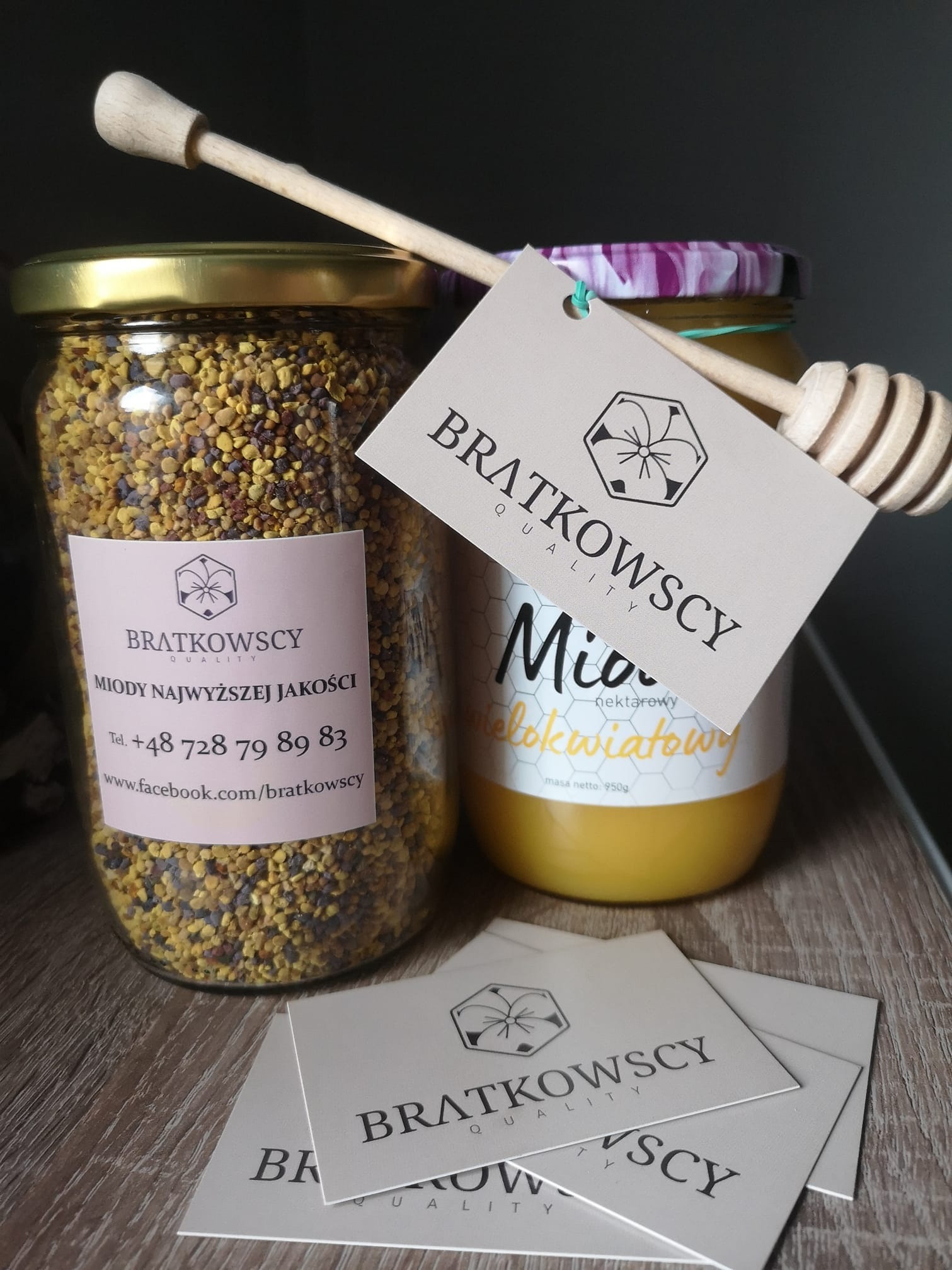 Dwa słoiki: jeden z pyłkiem pszczelim z etykietą, drugi z miodem wielokwiatowym, drewniany nabierak do miodu oparty o słoik, wizytówki firmy na drewnianym blacie.