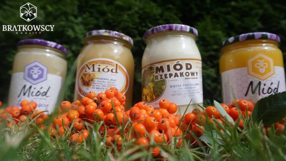 Cztery słoiki z różnymi rodzajami miodu, udekorowane owocami jarzębiny, ustawione na trawie, z widocznym logo producenta w lewym górnym rogu.