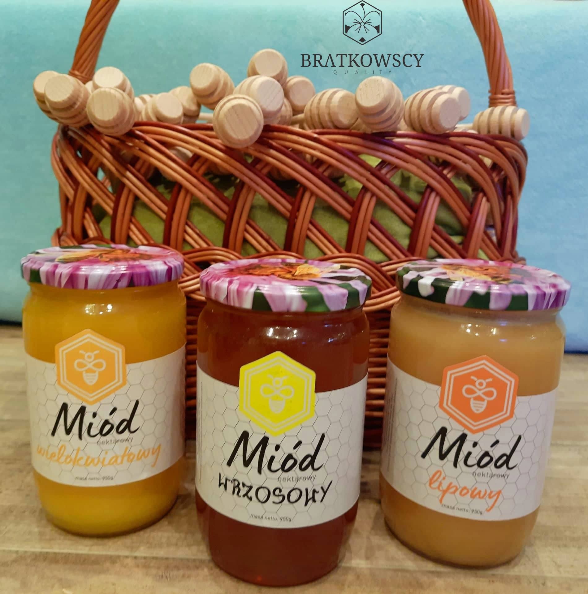 Trzy słoiki miodu z Szczecina: wielokwiatowy, wrzosowy i lipowy, ustawione przed wiklinowym koszem z drewnianymi nabierakami do miodu.