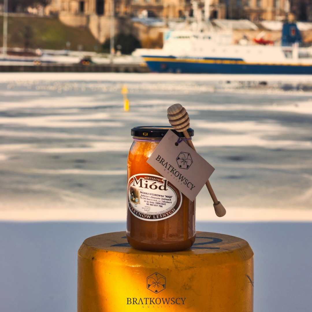 Słoik nektarowego miodu Bratkowscy z drewnianym nabierakiem i etykietą, ustawiony na żółtym słupku na tle zamarzniętego akwenu i statku.