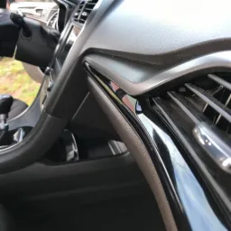 Wnętrze samochodu: deska rozdzielcza z kratką wentylacyjną i elementami z połyskliwego tworzywa sztucznego, widoczne drobne zabrudzenia na kratce.