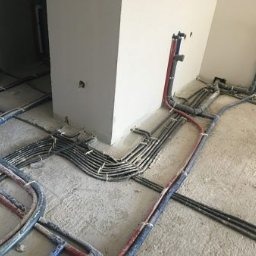 Rzędy rur instalacji wodnej i kanalizacyjnej ułożone na surowej posadzce w trakcie prac instalacyjnych, widoczne przyłącza do pionu i kolumny konstrukcyjnej.