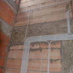 Wewnętrzna ściana z cegły, częściowo ocieplona, z widoczną instalacją elektryczną w białych rurkach PCV, układanych pionowo i poziomo.