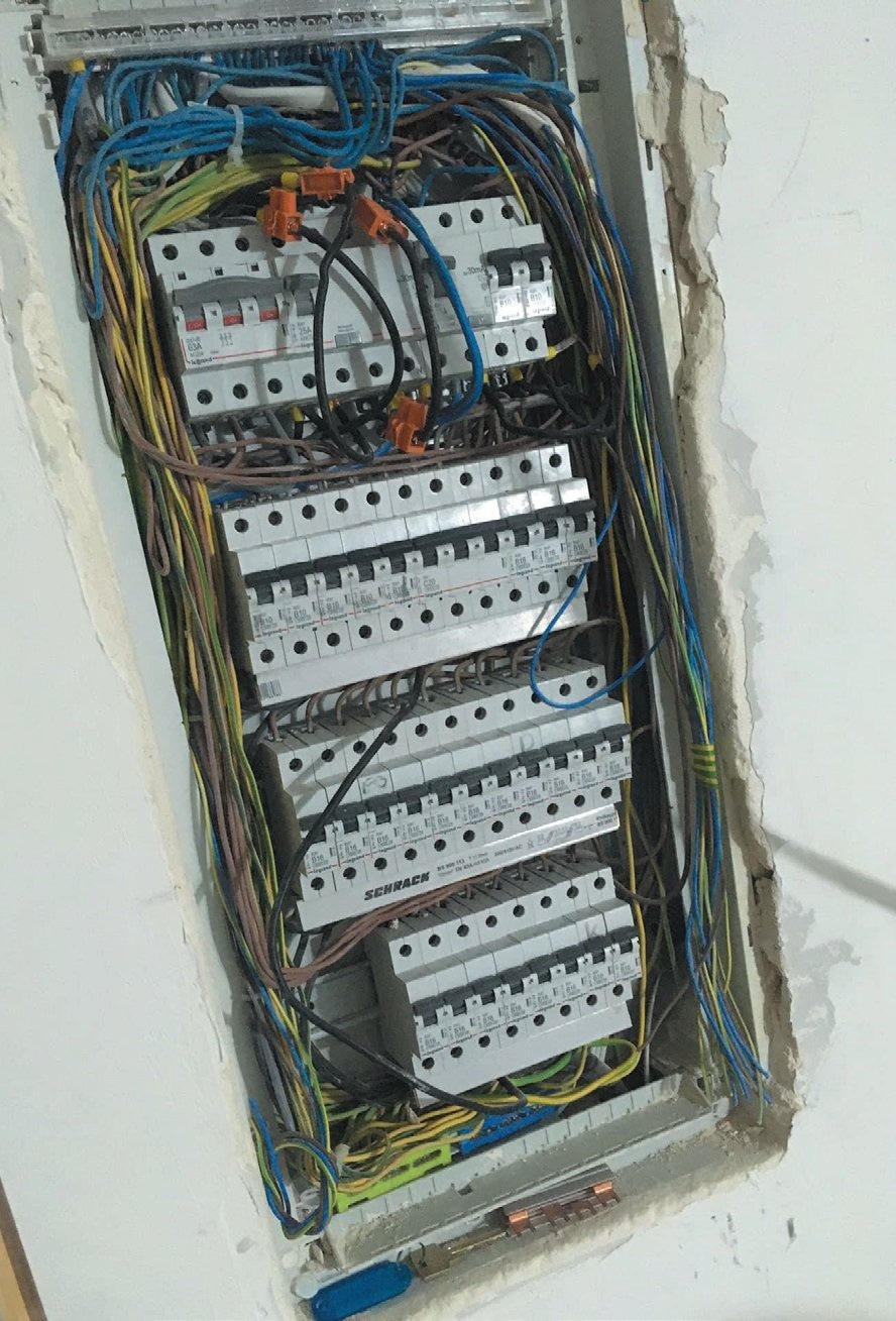 Rozdzielnica elektryczna w trakcie montażu, widoczne liczne przewody i zabezpieczenia, ściana z ubytkami tynku.