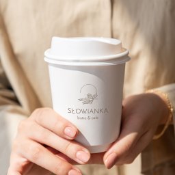 Kreowanie wizerunku marki - Anna Herman - Dłonie kobiety trzymają biały kubek z logo 'Słowianka bistro & cafe' w minimalistycznym stylu z motywem roślinnym.