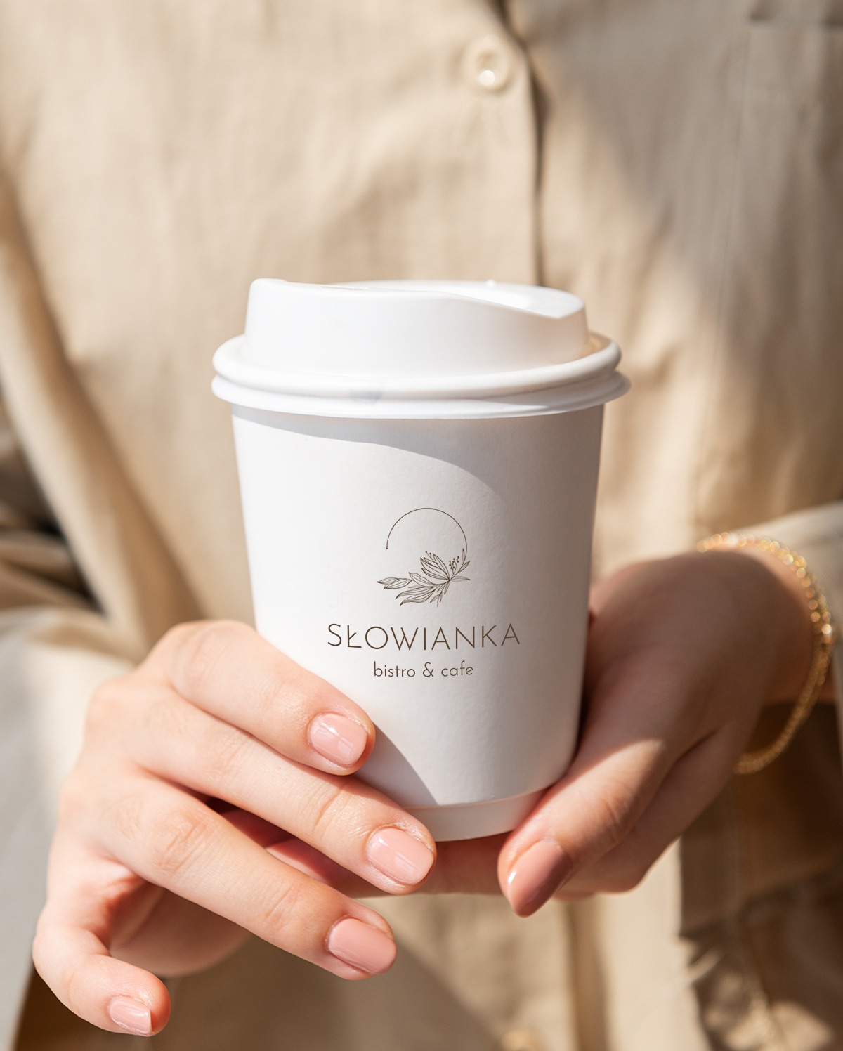 Dłonie kobiety trzymają biały kubek z logo 'Słowianka bistro & cafe' w minimalistycznym stylu z motywem roślinnym.