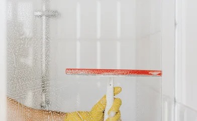 Osoba w żółtych rękawicach gumowych czyści szklaną kabinę prysznicową za pomocą ściągaczki z czerwoną gumą, usuwając pianę z mydlin.