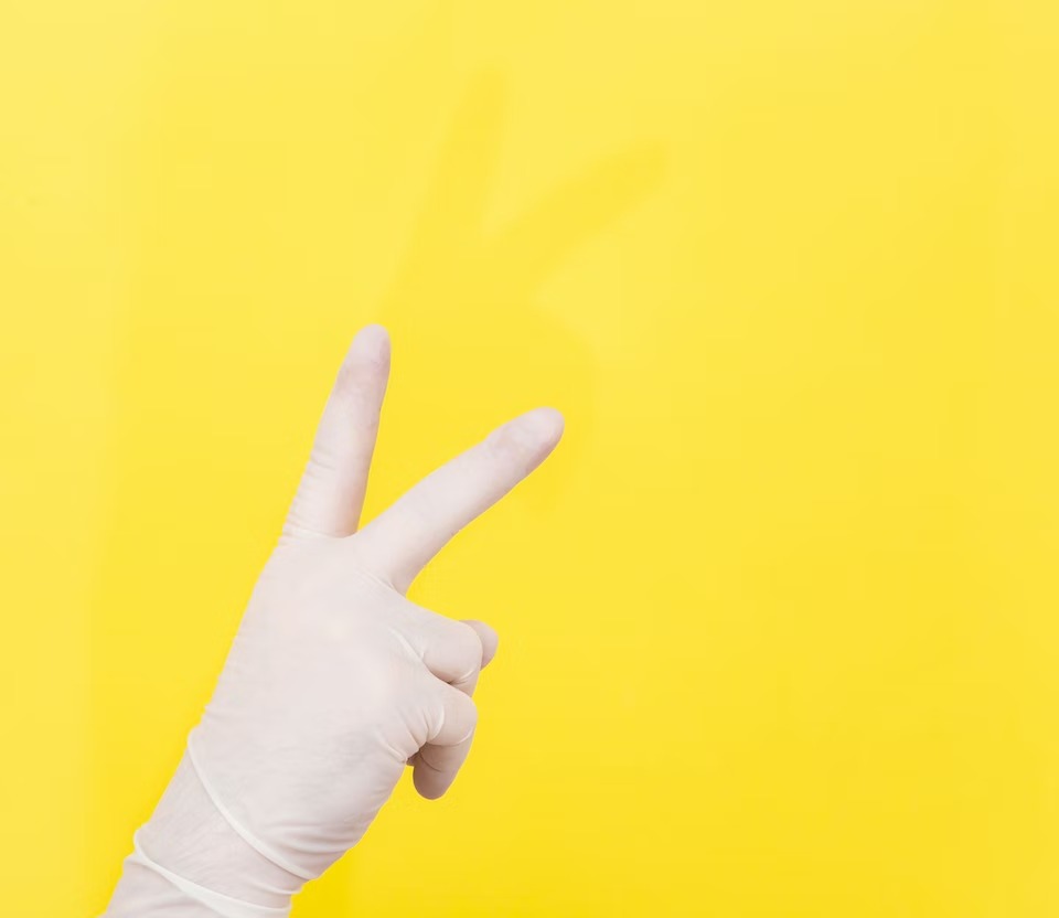 Dłoń w lateksowej rękawiczce pokazująca znak zwycięstwa na jaskrawożółtym tle, rzucająca delikatny cień.
