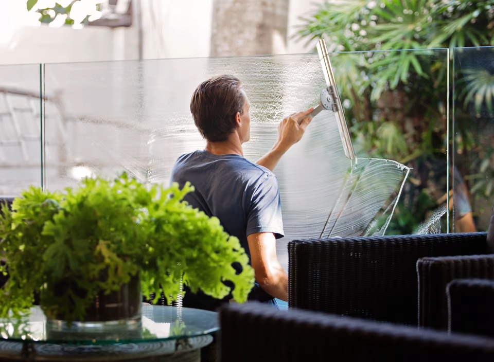 Mężczyzna w niebieskiej koszulce czyści szklaną balustradę balkonu za pomocą ściągaczki, widoczny efekt czyszczenia, w tle zieleń i meble ogrodowe.