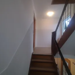 Wnętrze klatki schodowej z drewnianymi schodami, metalowymi balustradami i ścianą o nierównej fakturze.