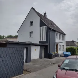 Dwukondygnacyjny dom jednorodzinny po renowacji elewacji, z szarą częścią boczną i białym frontem, widoczny z ulicy, z zaparkowanym czerwonym samochodem osobowym.