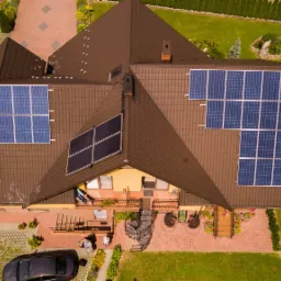 Widok z g&oacute;ry na dom z brązowym dachem i panelami słonecznymi; na posesji widoczny samoch&oacute;d, plac zabaw i spiralne schody.